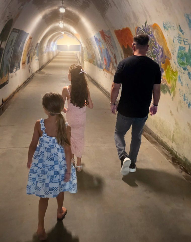 Steven and his two daughters, seen from behind, walking through a tunnel toward the beach at sunset
